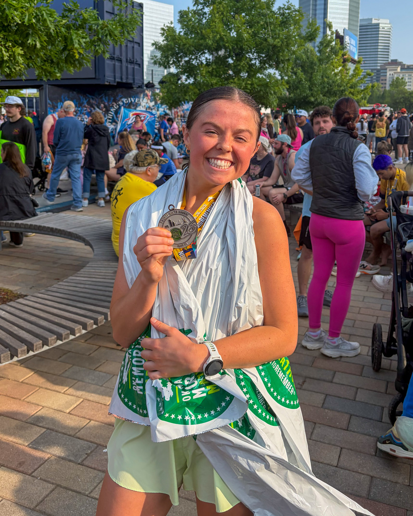 A smiling runner stands in a crowded downtown area holding up a marathon medal, wrapped in a foil blanket with other participants gathered behind her.