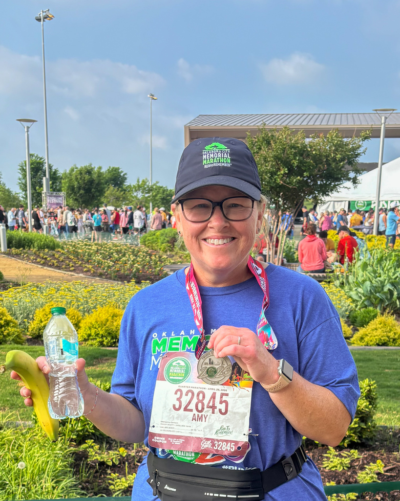 A runner wearing a hat and glasses smiles while holding a banana, water bottle, and medal, with a race bib visible and a garden area and crowd in the background.