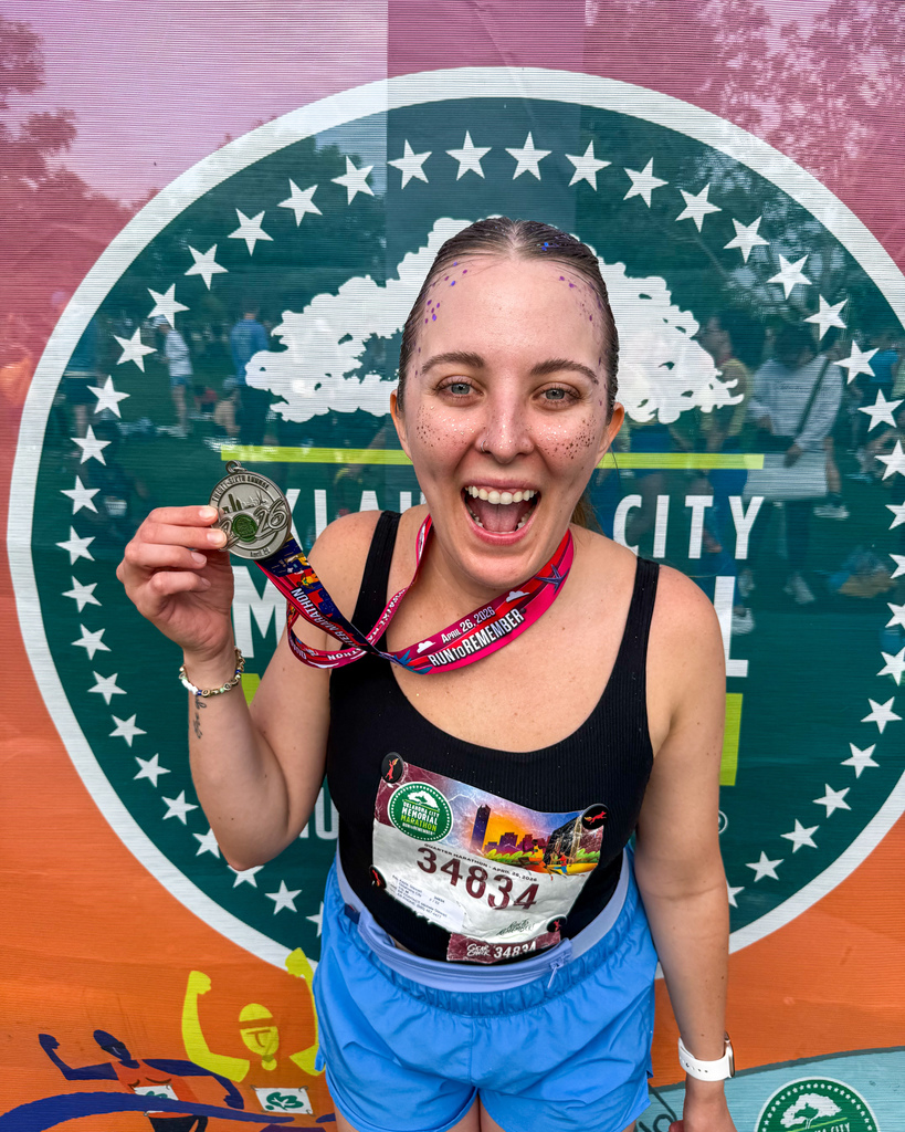A runner with face glitter smiles and holds up her medal in front of an Oklahoma City Memorial Marathon backdrop.