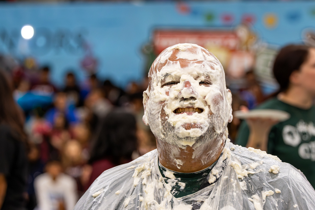 Staff member stands smiling with face fully covered in whipped cream, wearing a plastic poncho during a pie-in-the-face fundraising event.