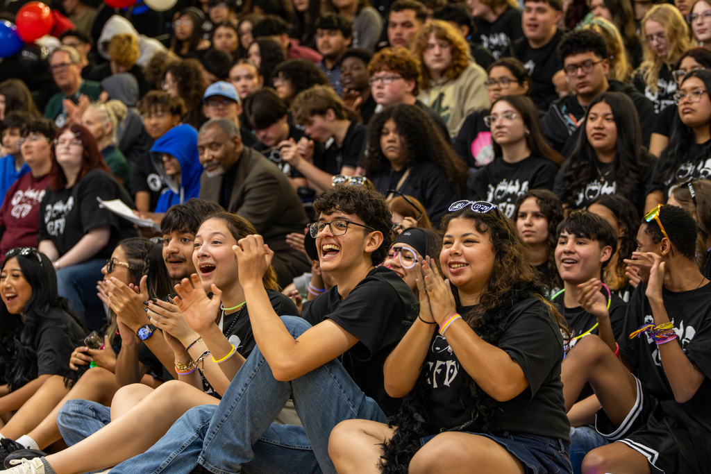 Packed gym crowd of students sits on the floor clapping and cheering during a school assembly.