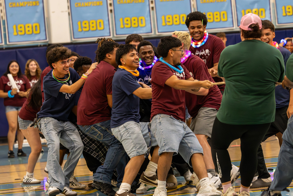 Group of students pulls together in a tug-of-war game in the gym while classmates cheer them on.