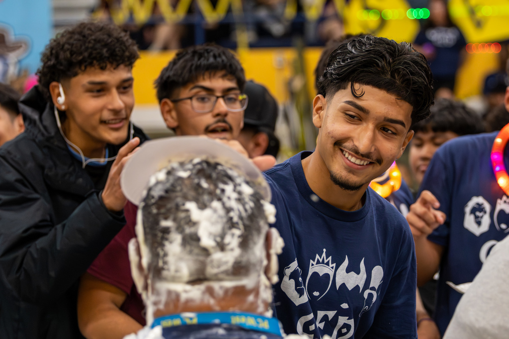 Students laugh as they dump a plate of whipped cream onto a staff member’s head during a school assembly fundraiser.