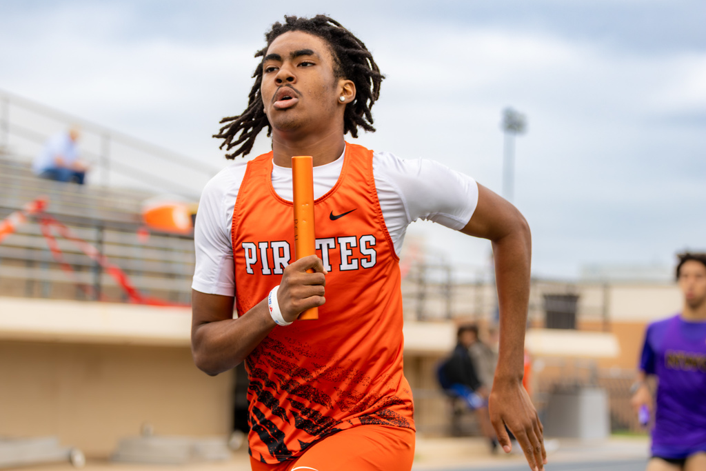 Male runner in an orange Pirates uniform sprints forward holding a relay baton during a race.
