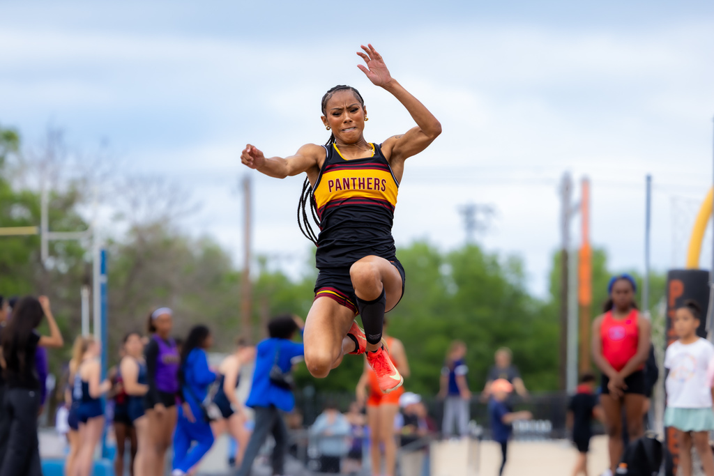 Female athlete in a Panthers uniform leaps into the air during a long jump attempt, arms raised for balance.