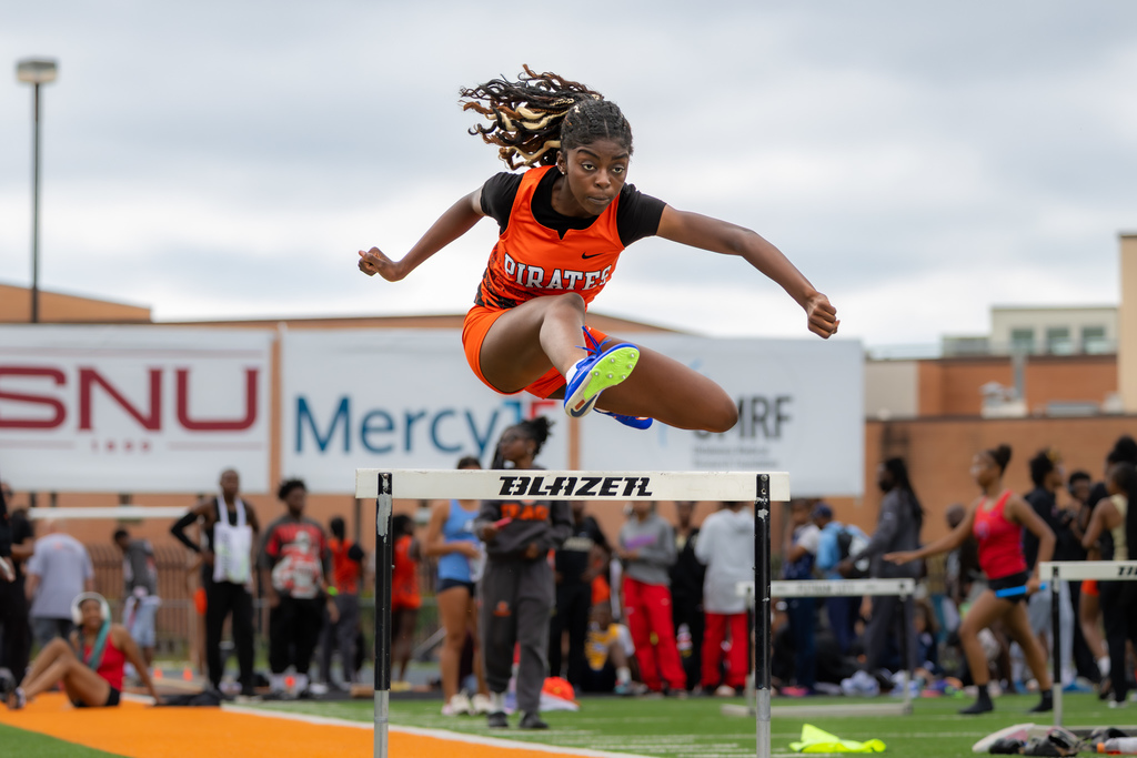 Female hurdler in an orange Pirates uniform clears a hurdle with legs extended and eyes focused ahead.