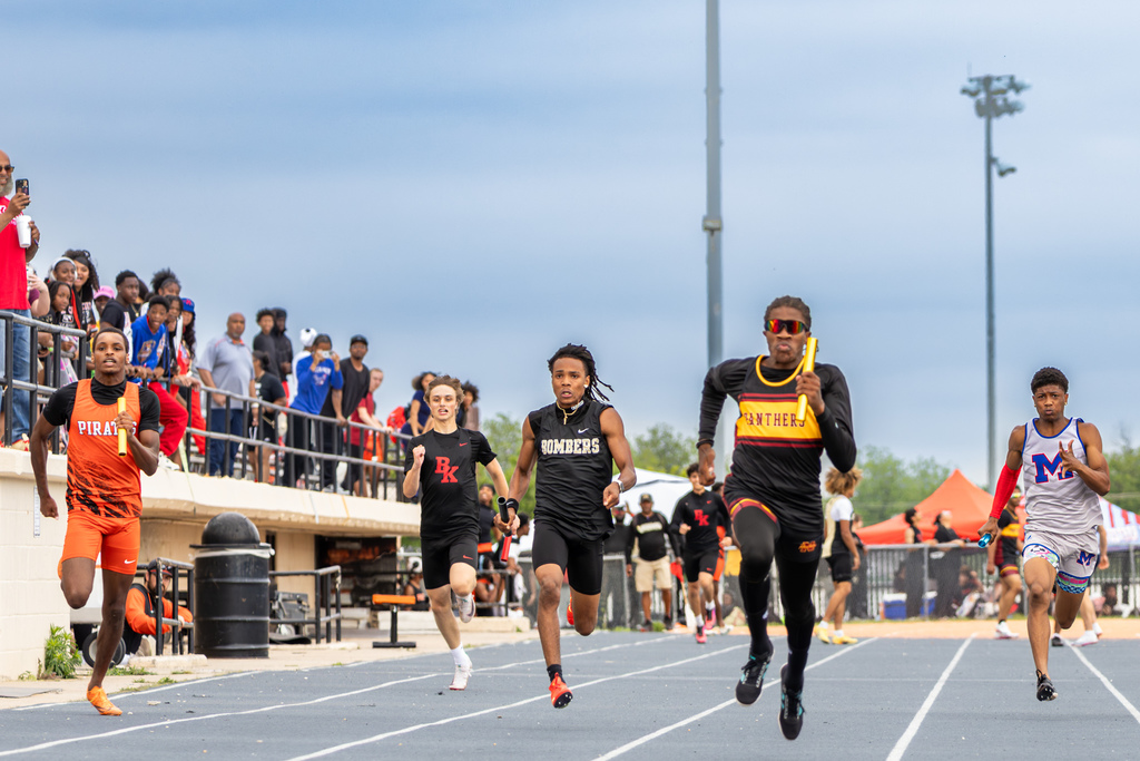 Relay runners sprint down the track toward the finish line, each holding a baton as spectators watch from the stands.