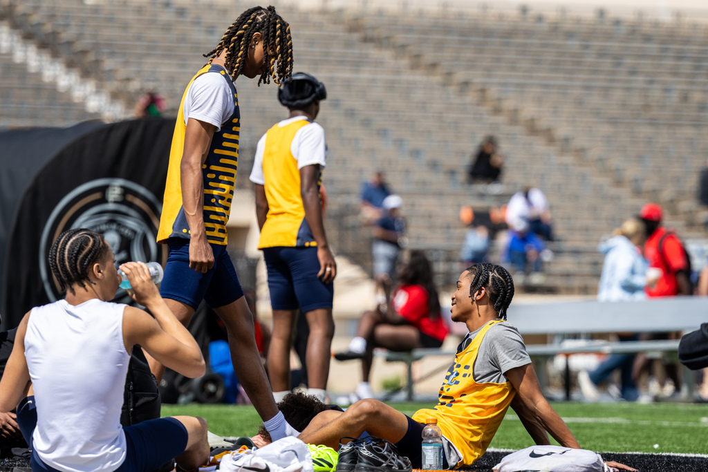 Track athletes in yellow uniforms rest and hydrate on the turf between events, sitting and standing near their gear.
