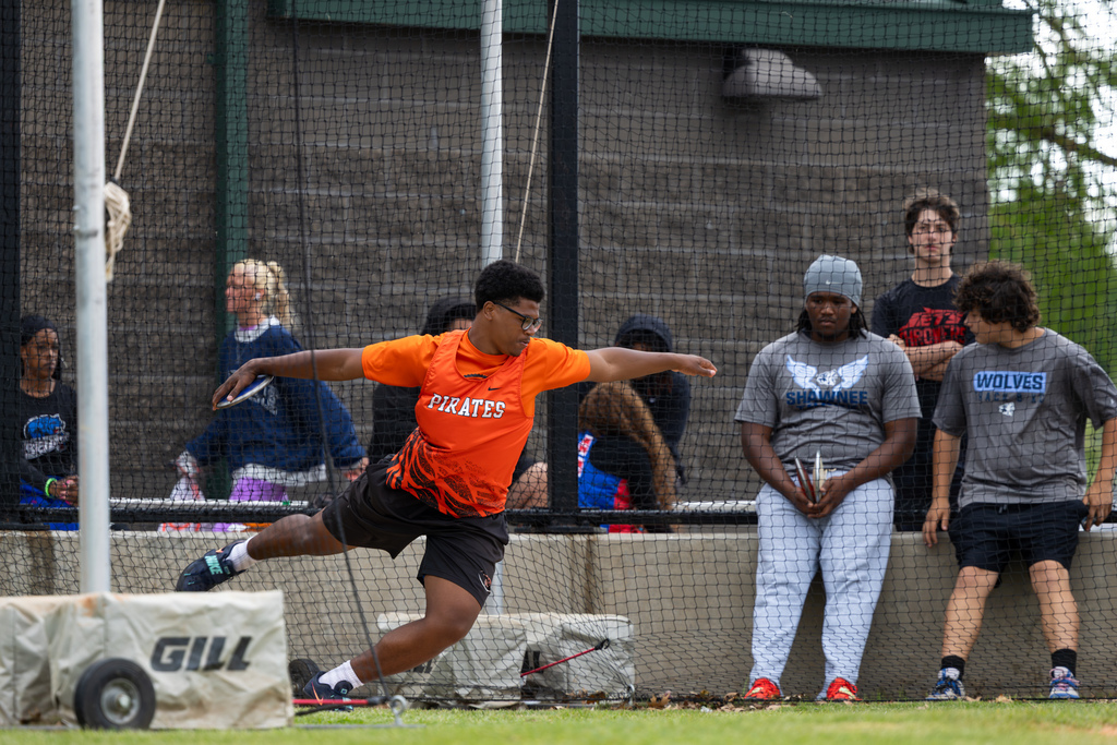 Male athlete in a Pirates uniform spins and releases a discus inside a netted throwing area while other competitors watch.