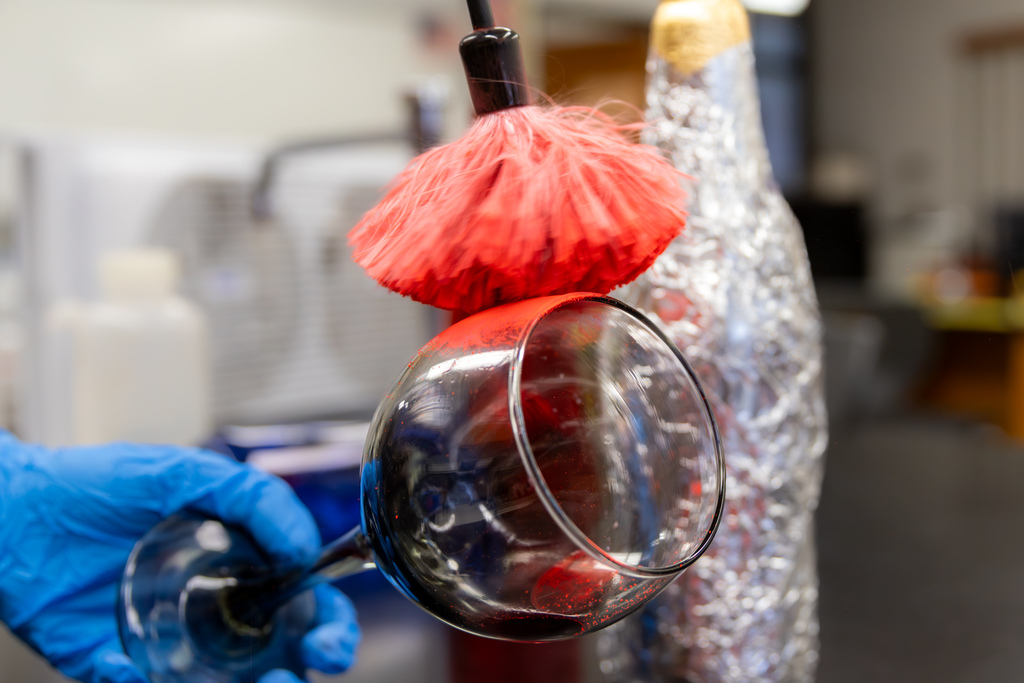 Close-up of a gloved hand dusting a glass with red fingerprint powder, with a foil-wrapped bottle in the background.