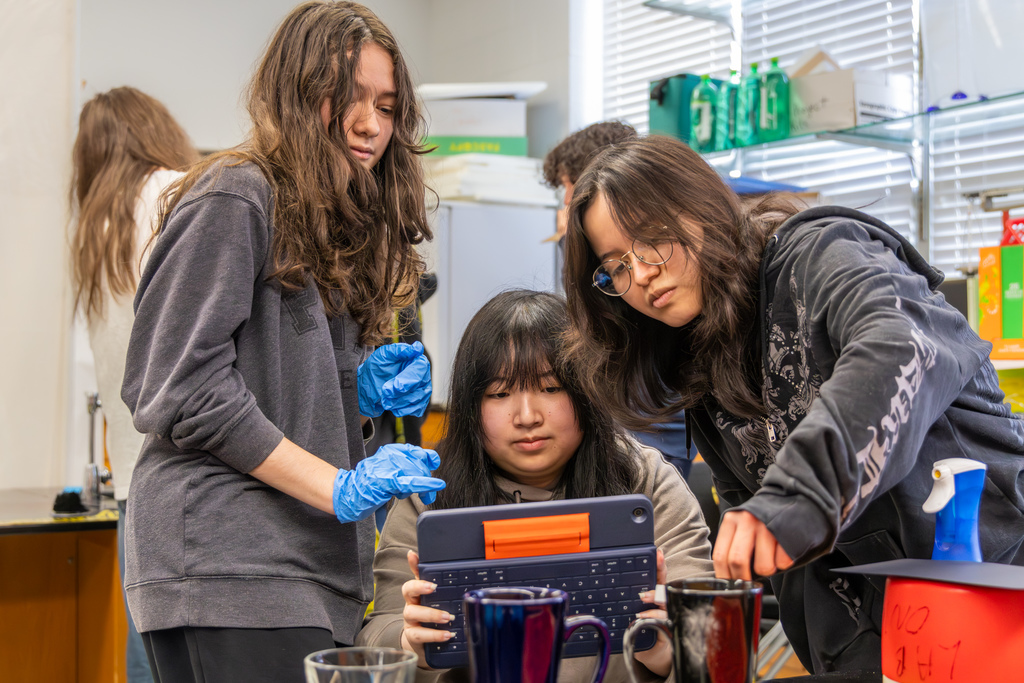 Three students collaborate at a lab table, reviewing evidence on a tablet while handling cups marked for investigation.
