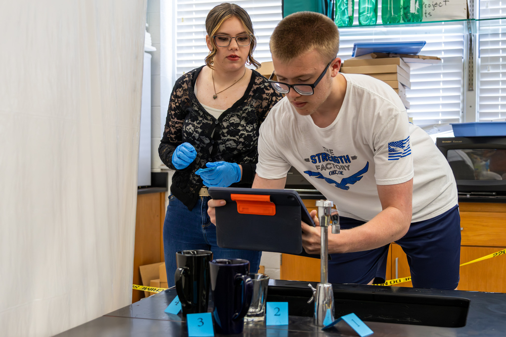Students examine a mock crime scene setup at a lab station, using a tablet to document evidence labeled around mugs and a sink.