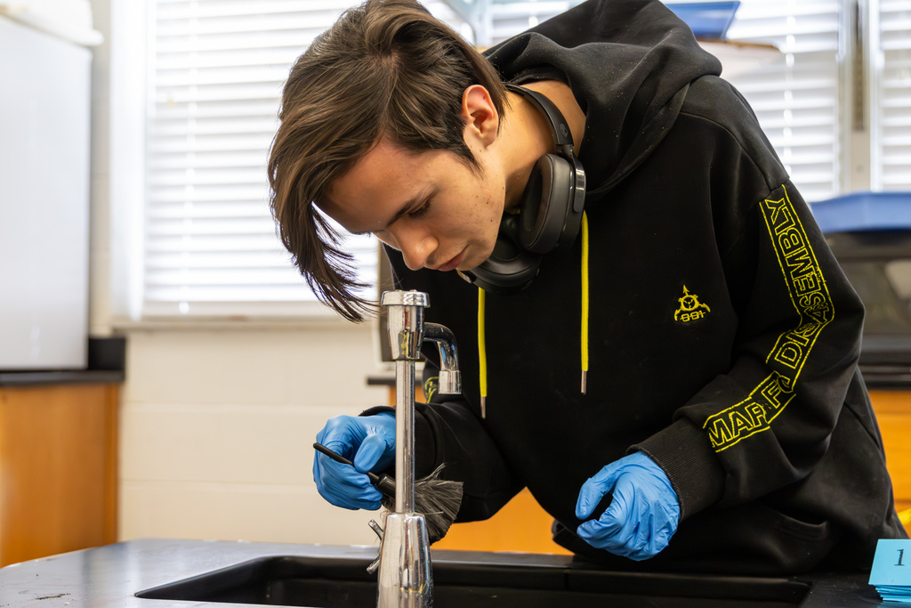 Student wearing gloves dusts a faucet for fingerprints using a brush during a forensic investigation activity.