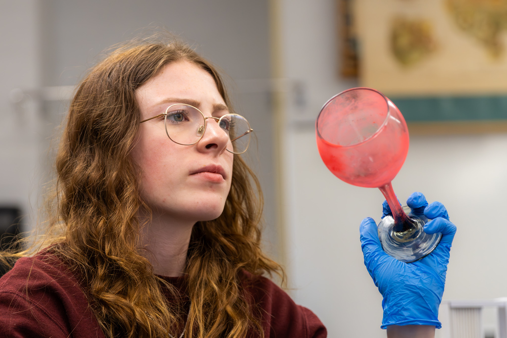 Student wearing gloves holds up a glass, inspecting red fingerprint powder for evidence.