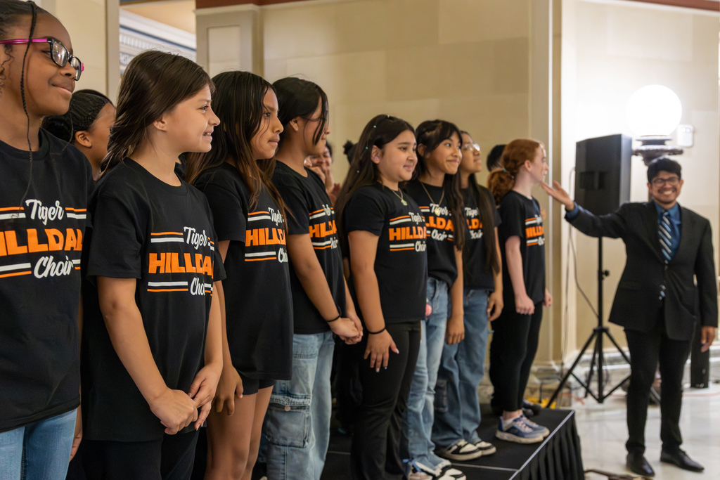 Side view of the choir lined up on stage as the conductor leads them through a song.