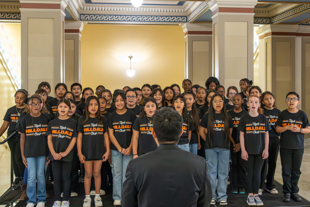 Front view of the choir singing together in matching black Hilldale Choir shirts as a conductor directs them.