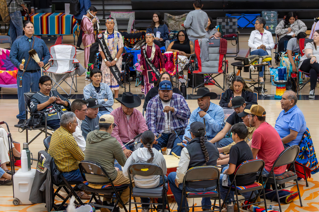A drum circle performs at the center of the Putnam City High School gym during the Putnam City Youth Powwow, with dancers and attendees gathered around them.