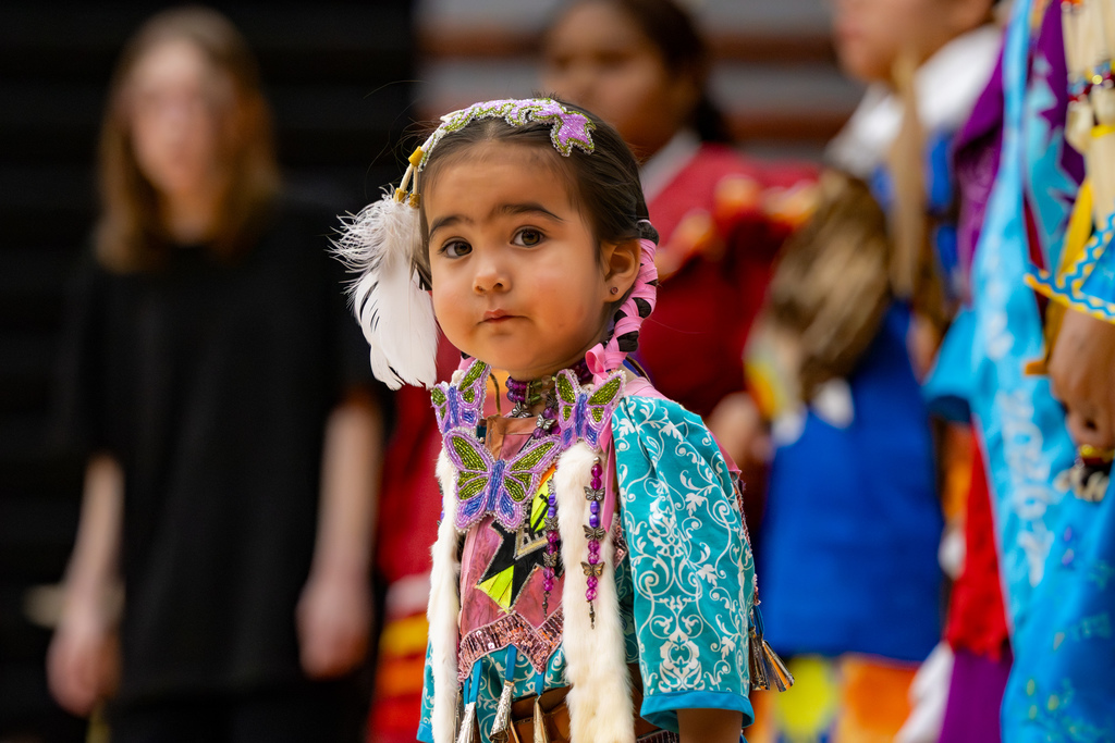 A young child in turquoise and pink regalia looks toward the camera while standing with other dancers during the Putnam City Youth Powwow.