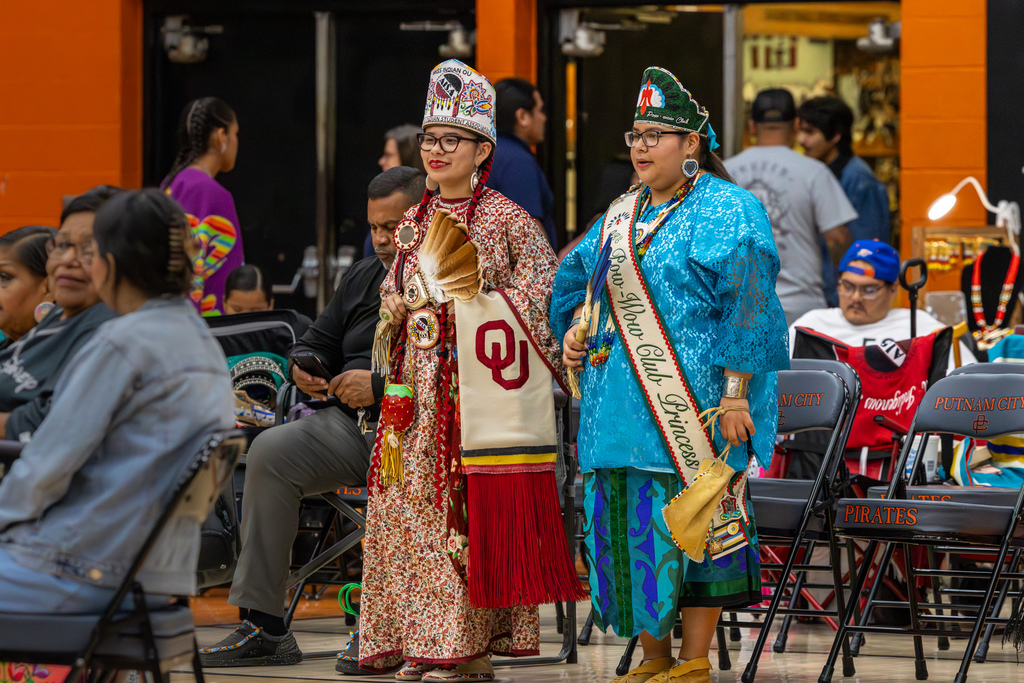 Two youth participants in traditional regalia stand together inside the Putnam City High School gym during the Putnam City Youth Powwow, surrounded by seated attendees and folding chairs.