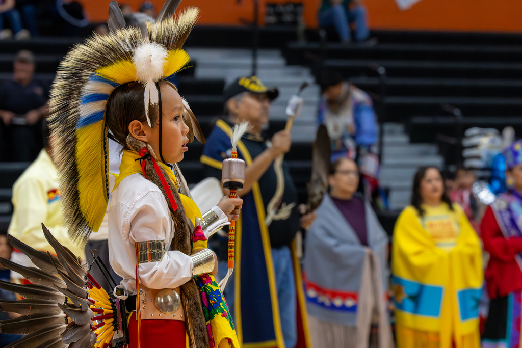 A young dancer in bright yellow, blue, and red regalia stands in profile while holding a decorated dance staff during the Putnam City Youth Powwow.