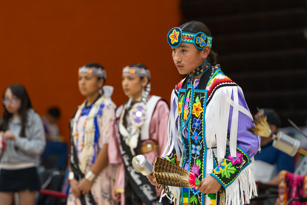 A young dancer in colorful traditional regalia stands with other youth dancers lined up behind him in the Putnam City High School gym.
