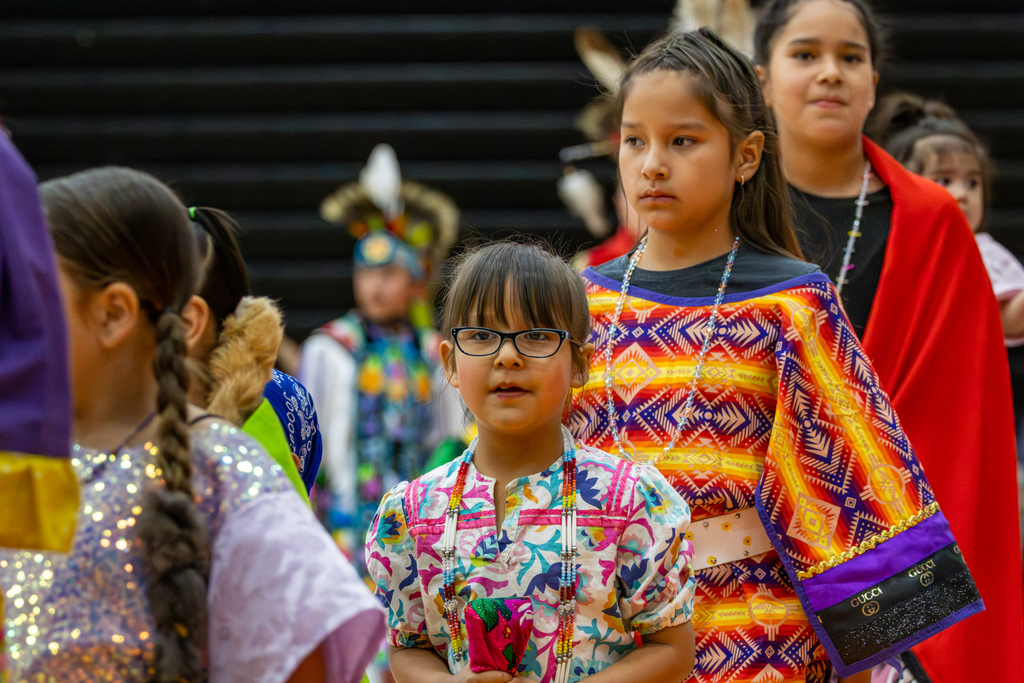 Several young dancers in bright patterned regalia stand together during the Putnam City Youth Powwow, with one child looking toward the camera.