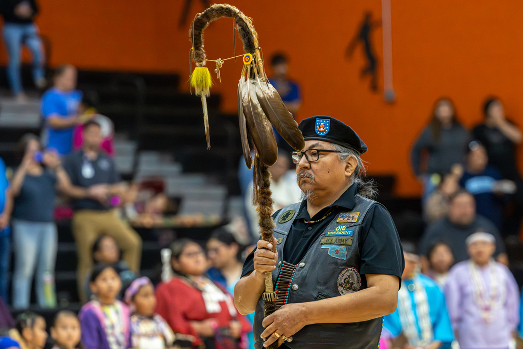 An adult participant holds a decorated staff during the Putnam City Youth Powwow, with dancers and attendees visible in the background.