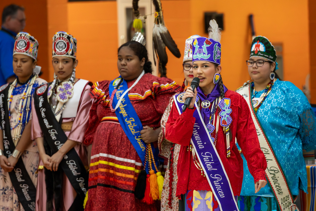 Youth royalty and other participants in traditional regalia stand together as one student speaks into a microphone during the Putnam City Youth Powwow.
