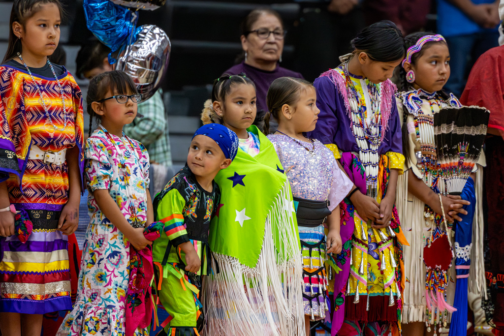 A group of young children in colorful regalia stand together during the Putnam City Youth Powwow inside the Putnam City High School gym.