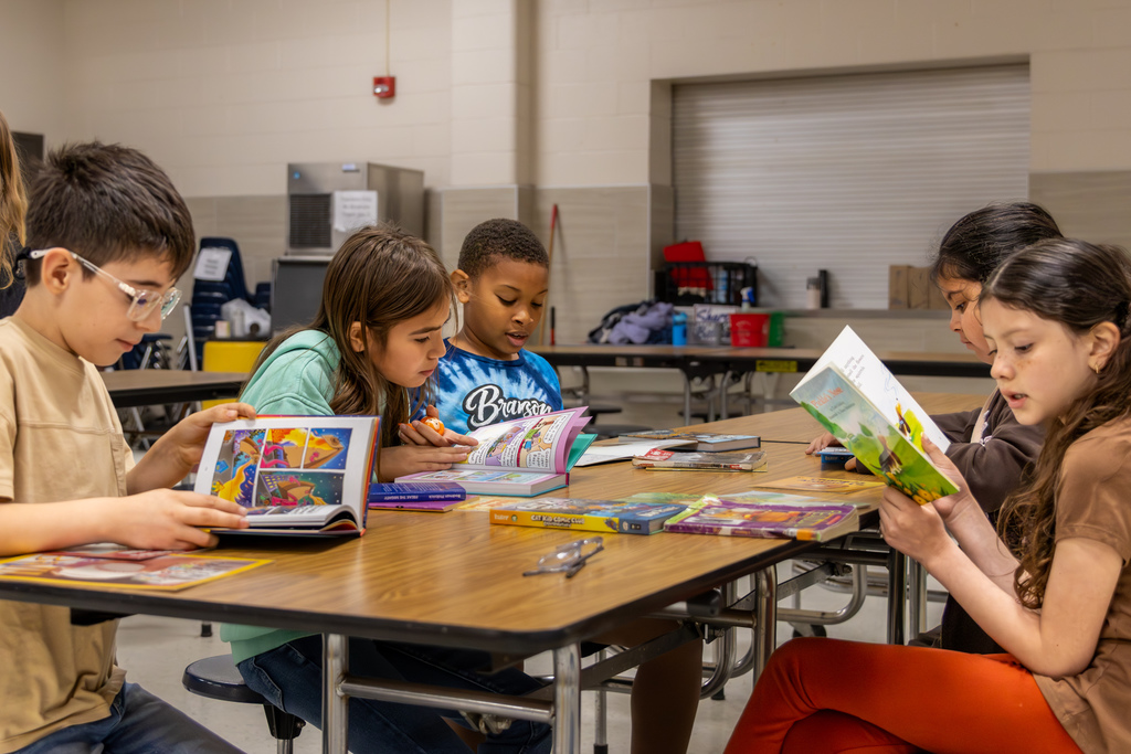 A group of Overholser Elementary students sit together at a cafeteria table, reading and looking through books from the Free Book Fair.