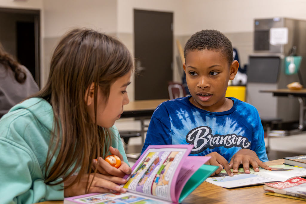 Two students sit side by side at a table, looking through books and talking during Overholser Elementary’s Free Book Fair.