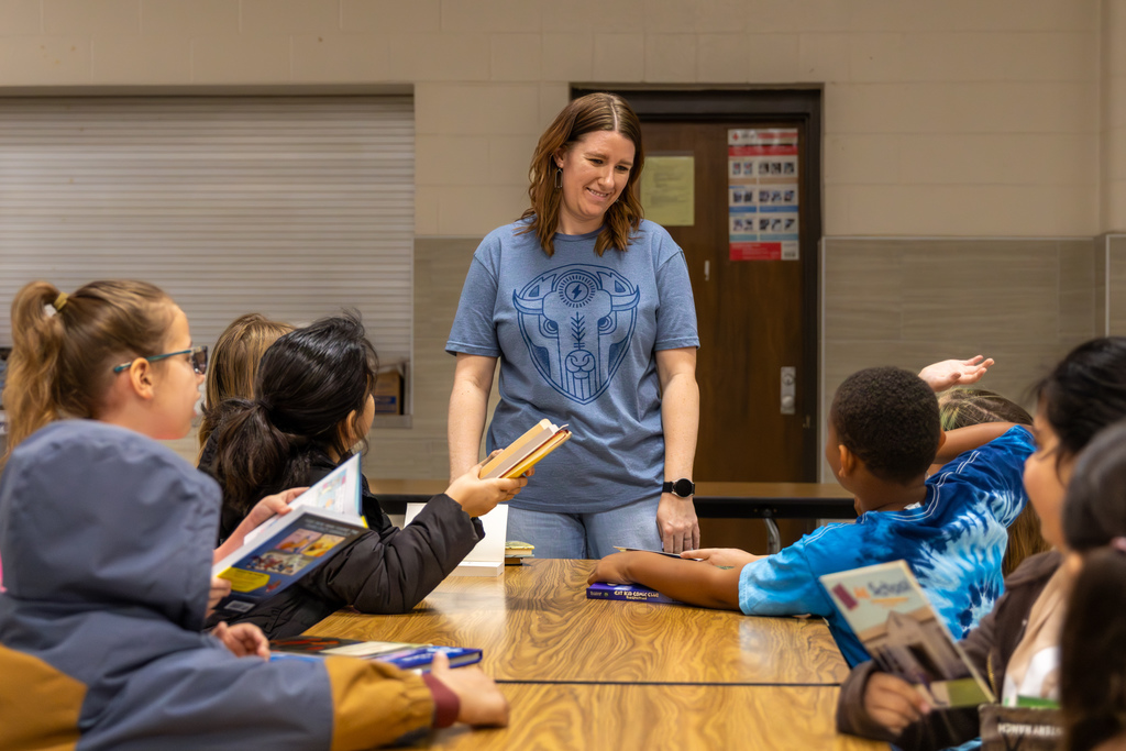 A staff member smiles while students ask questions and hold books around a cafeteria table at Overholser Elementary’s Free Book Fair.