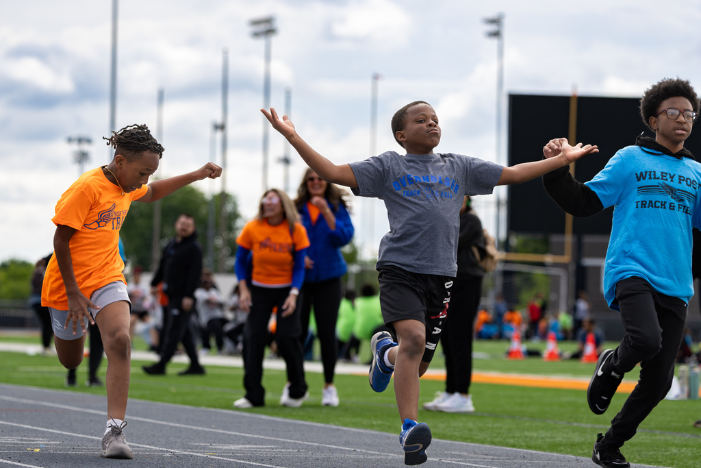 Elementary students run down the track during a race, with school staff and spectators in the background.