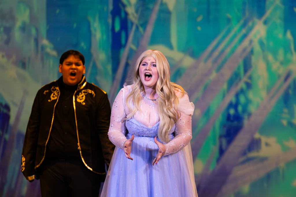 Putnam City North students perform a vocal scene from Frozen: The Broadway Musical, with Elsa singing at center stage in front of an icy projected backdrop.