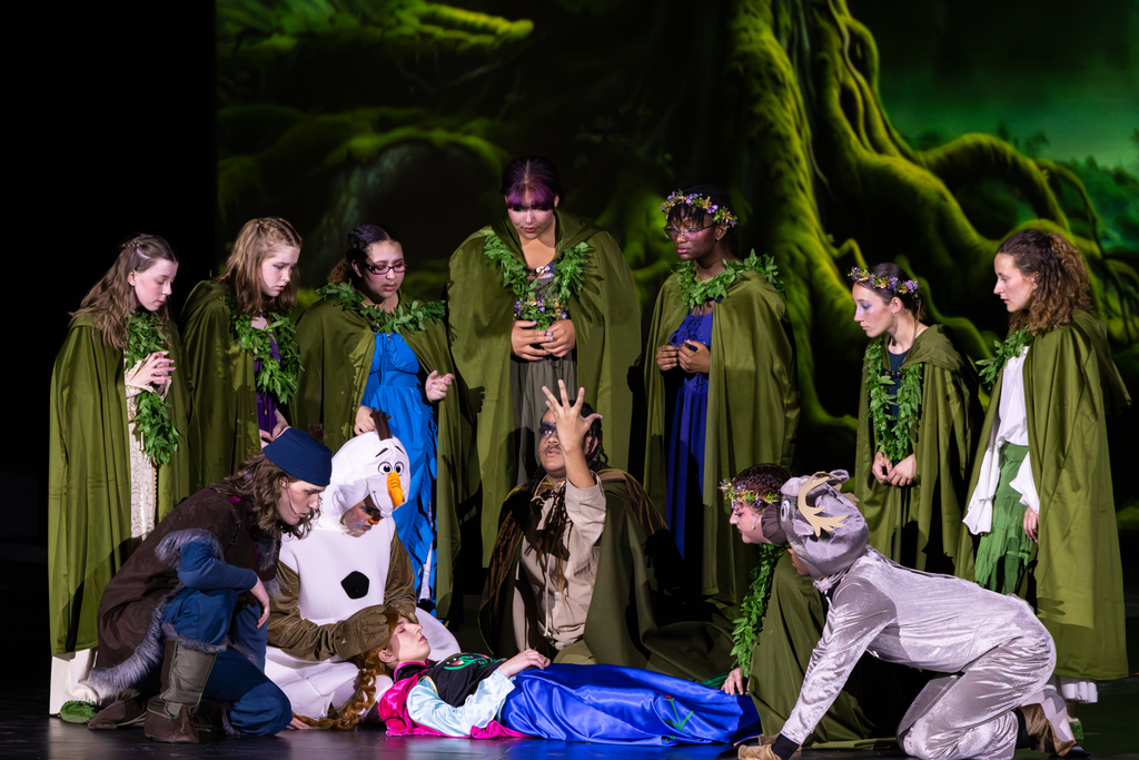 Putnam City North students perform a dramatic scene from Frozen: The Broadway Musical, with ensemble members gathered around Anna as she lies on stage.