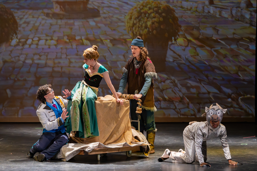 Putnam City North students perform a scene from Frozen: The Broadway Musical, with students in royal and animal costumes gathered around a covered cart on stage.