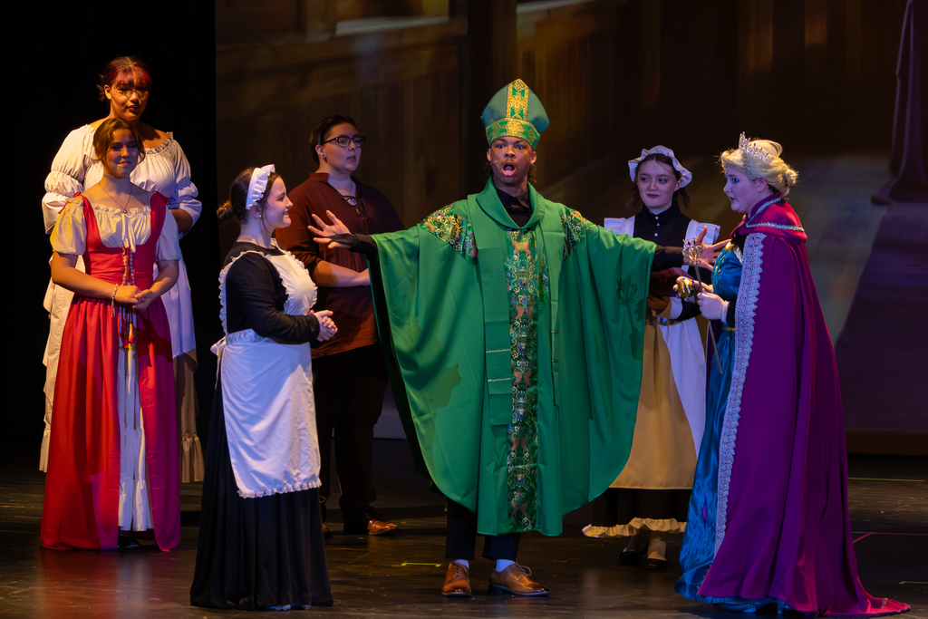 Putnam City North students perform a castle scene from Frozen: The Broadway Musical, with one student in a green ceremonial robe standing center stage among other cast members in period costumes.