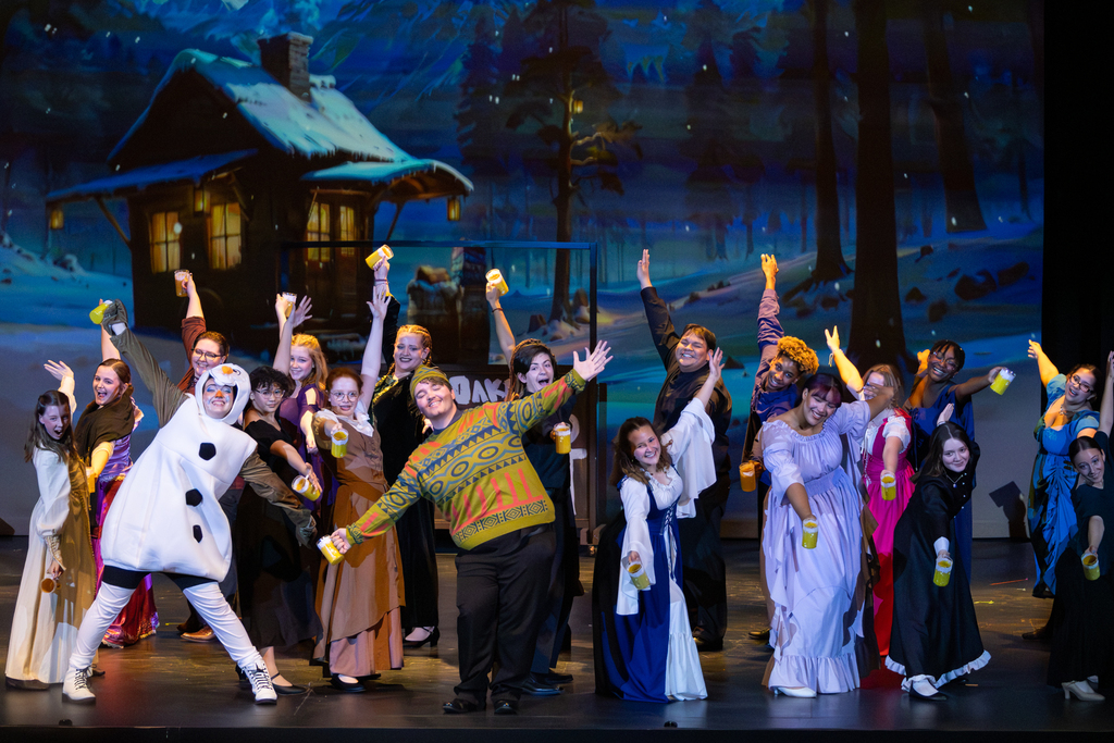 Putnam City North students perform a lively ensemble number from Frozen: The Broadway Musical, posing with raised arms and cups in front of a snowy cabin backdrop.