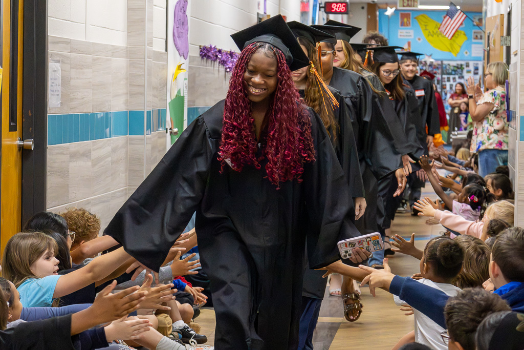 Putnam City seniors in black caps and gowns walk through an elementary hallway as younger students line the floor and reach out for high-fives.