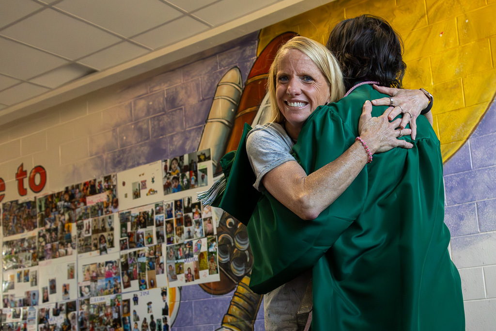 A Putnam City senior in a green cap and gown hugs a former teacher in front of a school memory wall.
