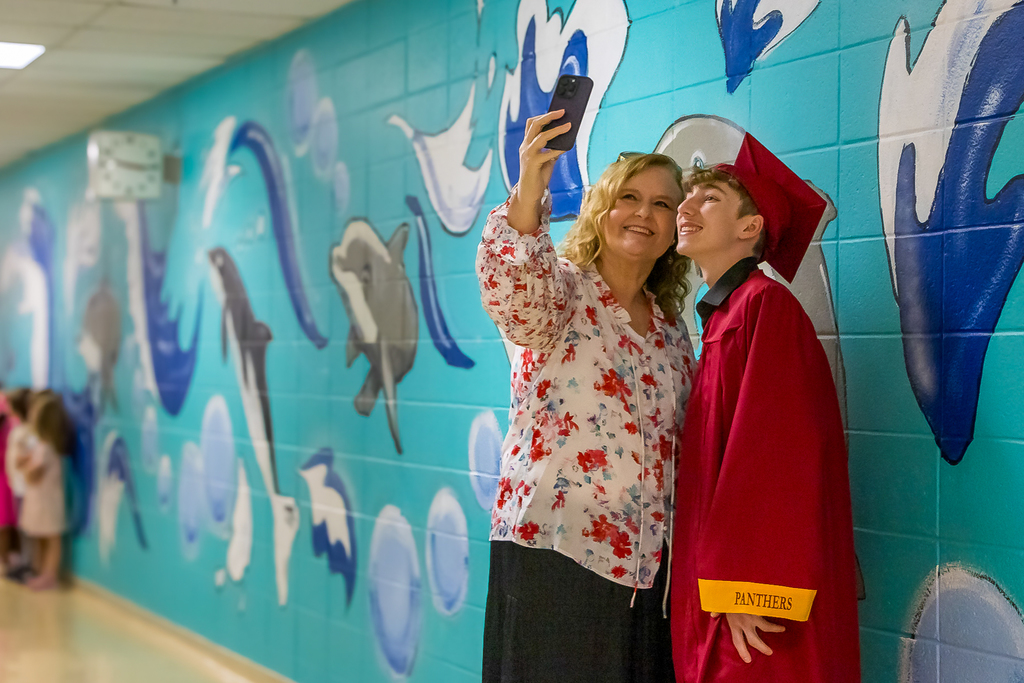 A Putnam City senior in a red cap and gown takes a selfie with an elementary teacher in front of a dolphin mural during a Grad Walk.