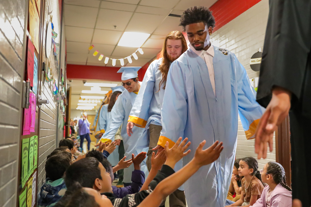 Putnam City seniors in light blue caps and gowns walk through an elementary hallway as younger students reach out for high-fives.