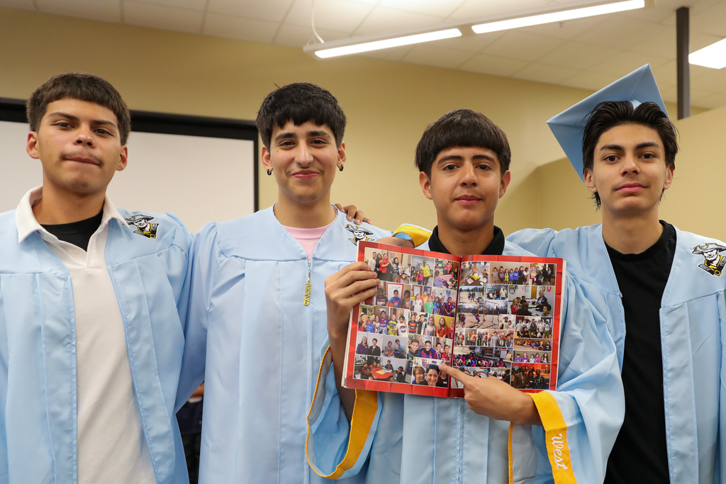 Four Putnam City seniors in light blue caps and gowns pose together while holding open a school memory book.