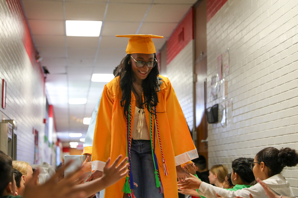 A Putnam City senior in a gold cap and gown walks through an elementary hallway as younger students reach out for high-fives.