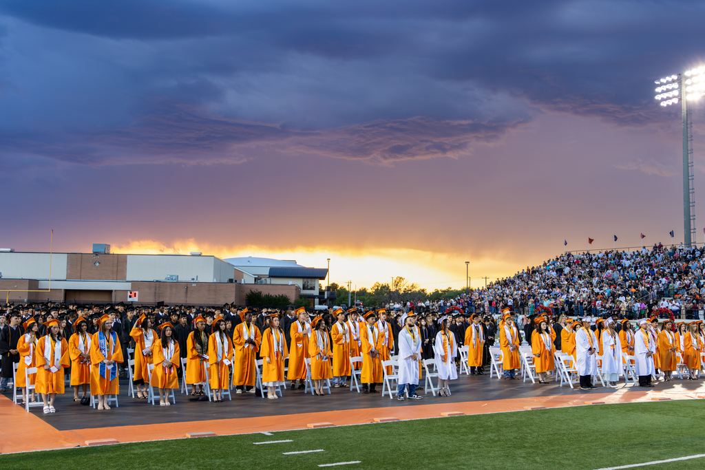 A wide group of students in graduation gowns standing on a football field with storm clouds in the background