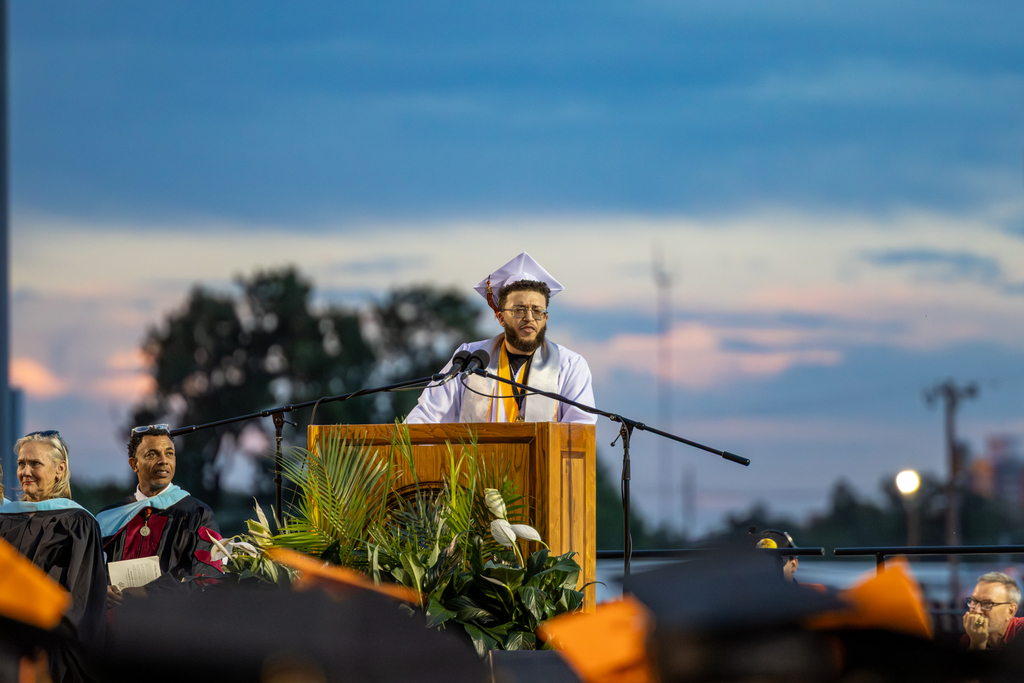 A student in a white graduation gown delivers a speech to a seated audience with storm clouds visible behind
