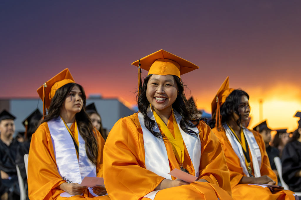 A student in a gold gown smiles at the camera with a golden sunset in the background