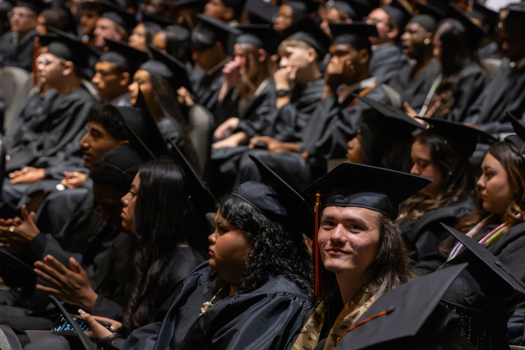 One student in a black graduation gown smiles at the camera in a sea of other graduates