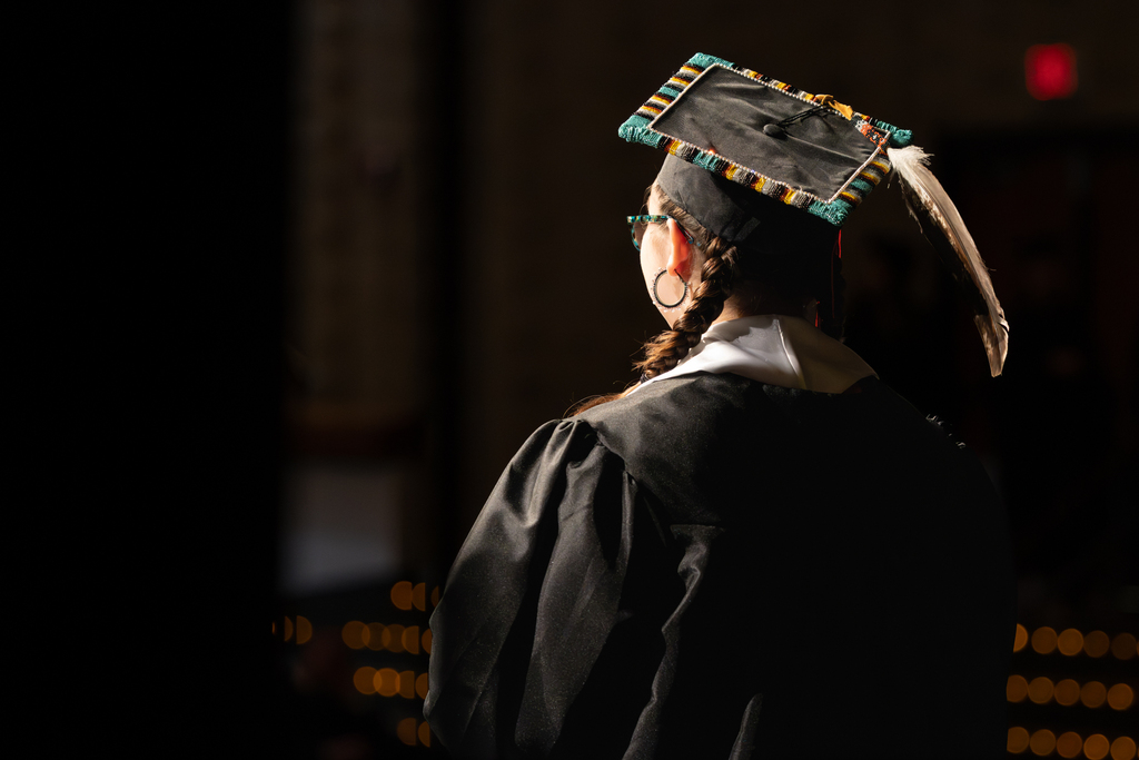 A student looks out across the stage, wearing a graduation cap decorated with symbols of heritage