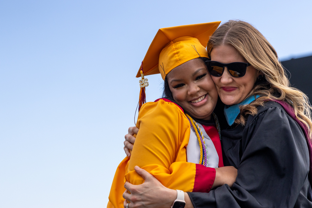 A Putnam City North graduate in a gold cap and gown hugs a staff member in academic regalia during graduation.